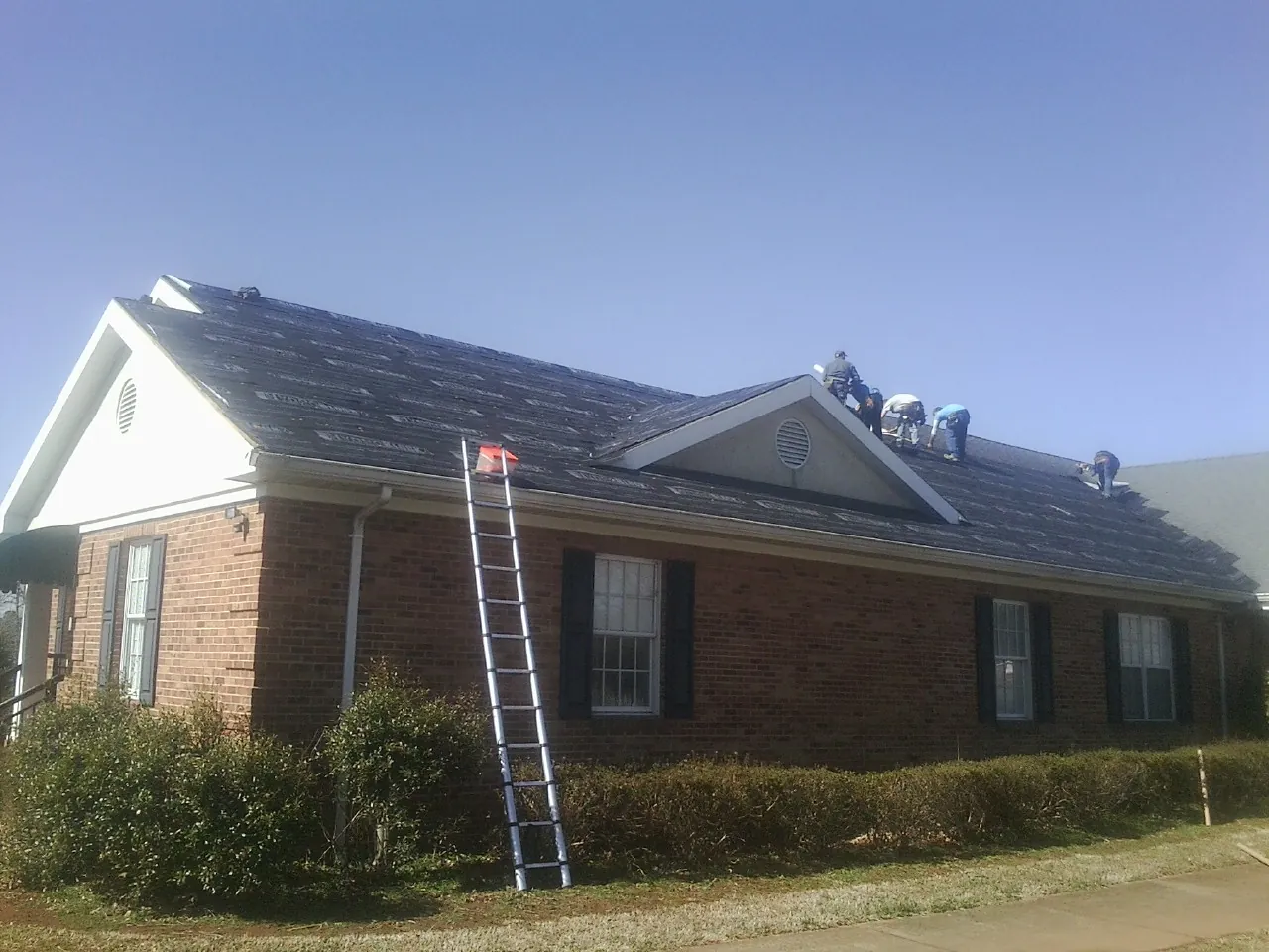 Crew tearing shingles off a brick ranch roof with a ladder set — Metal Roof Repair work by our crew in Enoree