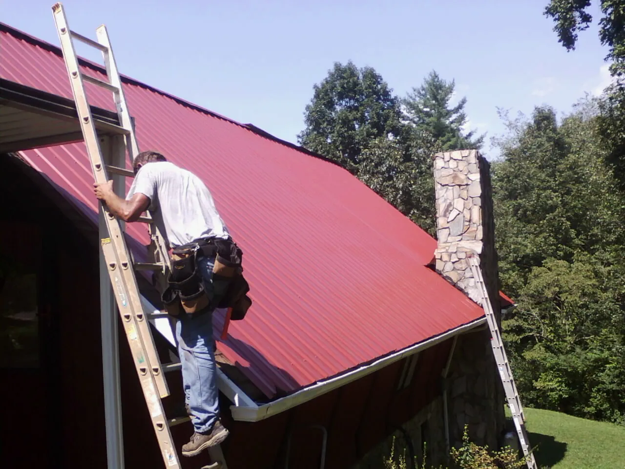 Roofer on a ladder beside a red metal roof and stone chimney — Soffit & Fascia Repair by Roofing Brothers in Enoree